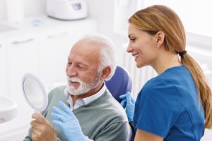 a senior man in the dental chair looking at himself in the mirror with a dental assistant looking on, illustrating a full-mouth reconstruction